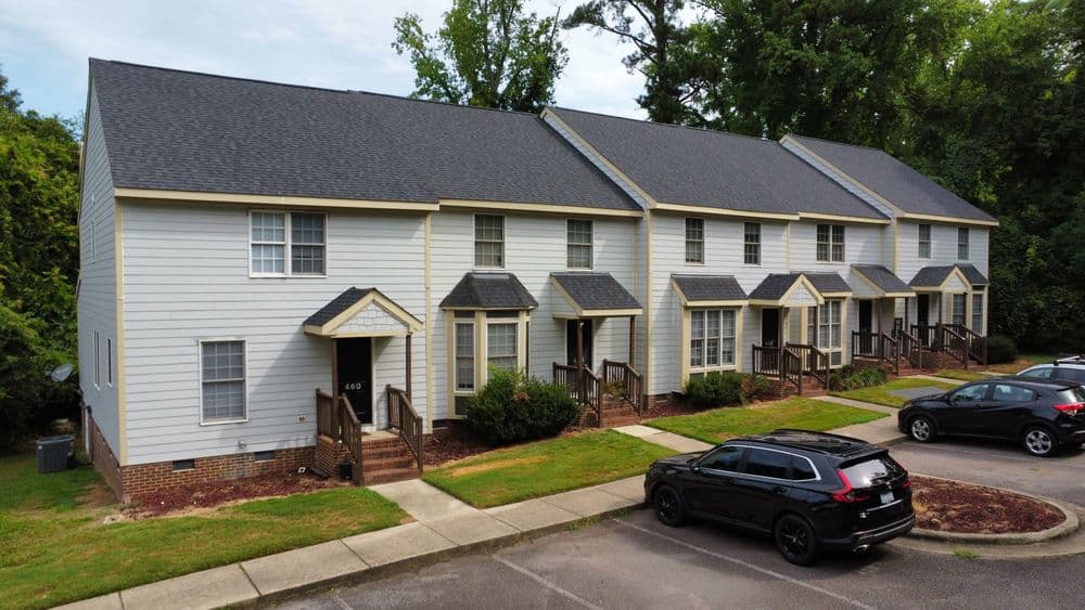 Row of townhouses with gray roofs and paved parking lot, surrounded by greenery.