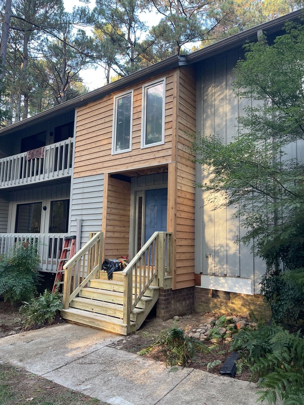 Newly renovated apartment entrance with wooden steps and modern siding in a wooded area.
