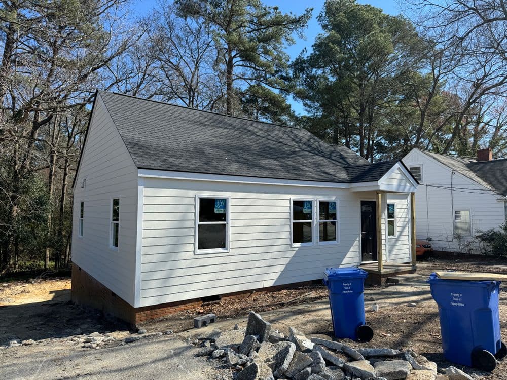 Newly renovated white house with black roof, surrounded by trees and blue trash bins.